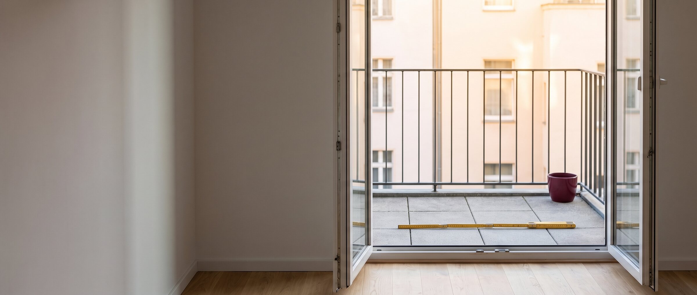 KI-generiertes Symbolbild: Blick aus einem leeren Zimmer durch die offene Balkont&uuml;r auf einen Balkon mit Metallgel&auml;nder, auf dem Boden liegt ein gelber Zollstock